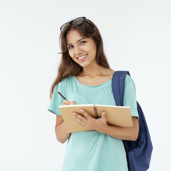 Picture of cheerful positive Hispanic college girl with eyeglasses on her head and bag on one shoulder posing in studio, smiling happily while writing something down in her copybook or diary